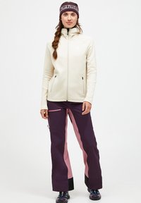 Light cream zip-up jacket with high collar, paired with dark purple pants featuring pink side panels and zippered pockets. Textured fabric.