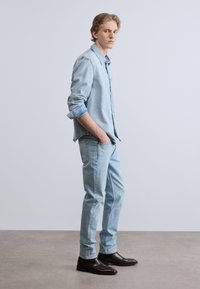 Ανοιχτό γαλάζιο πουκάμισο από τζιν με ρυμουλκούμενα μανίκια, σε συνδυασμό με ανοιχτό γαλάζιο τζιν παντελόνι και σκούρα καφέ μποτάκια αστραγάλου, τοποθετημένα σε ουδέτερο φόντο.