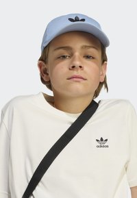 Casquette de baseball bleu clair avec logo noir brodé, portée avec une chemise blanche à manches courtes arborant un petit logo noir, et une bandoulière noire.