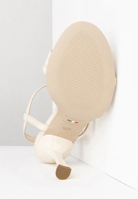 Beige hoge hakken sandaal met een glanzende afwerking, voorzien van slanke enkelbandjes en een getextureerde zool. De hoogte van de hak is visueel opvallend.