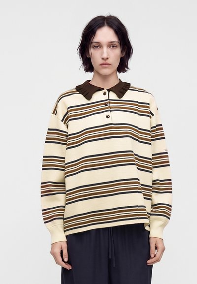 Jeune personne aux cheveux courts et foncés portant un pull crème avec des rayures horizontales marron et noir et un col marron foncé sur un fond uni.