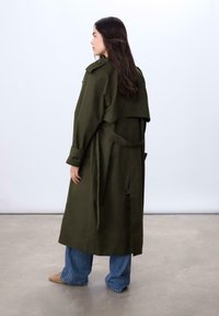 Olivengrøn trenchcoat med dobbeltbrystet design, oversized krave og bælte. Tætsiddende ærmer og løs pasform, kombineret med blå vide jeans.