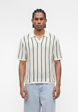 Junger Mann mit geflochtenem Haar, der ein weißes, kurzärmeliges Strick-Poloshirt mit vertikalen grünen Streifen und hellblauer Jeans trägt.