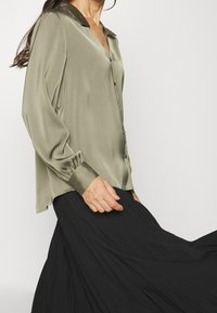 Blouse en satin vert olive avec un col sombre, dotée d'une fermeture à boutons sur le devant et de manches froncées, associée à une jupe noire fluide.