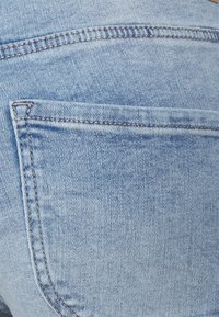 Jeans de mezclilla azul claro con un bolsillo trasero con costuras visibles y una textura ligeramente desgastada. La tela tiene un patrón de tejido clásico.
