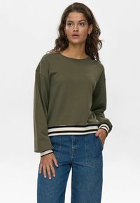 Felpa verde oliva cropped con spalle scese, collo a coste e polsini a righe. Abbinata a jeans a gamba larga blu.