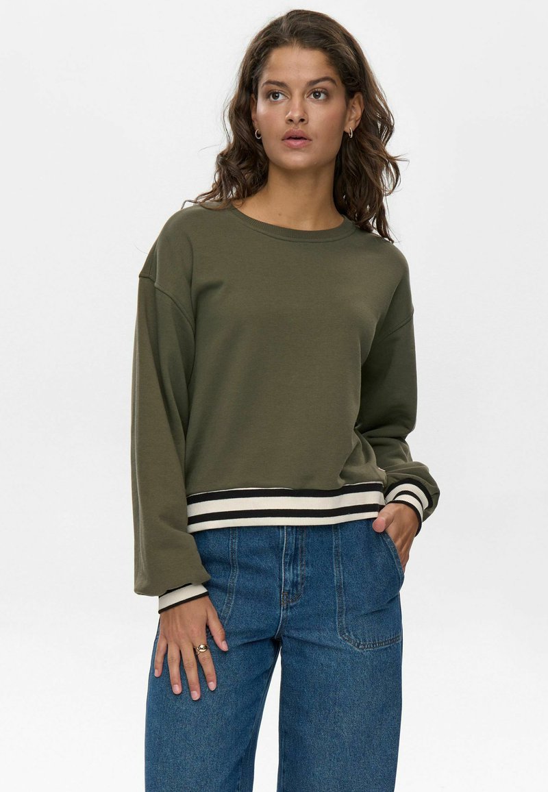 Felpa verde oliva cropped con spalle scese, collo a coste e polsini a righe. Abbinata a jeans a gamba larga blu.