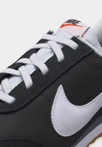 Μαύρο και άσπρο αθλητικό παπούτσι Nike με λευκά κορδόνια, μεγάλο λευκό λογότυπο swoosh και κόκκινο κείμενο «NIKE» στη γλώσσα, πάνω σε απλό φόντο.