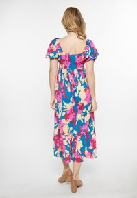 Blumiges Maxikleid in lebhaftem Blau, Pink und Gelb; puffige Ärmel; quadratischer Rückenausschnitt; gestuftes Saumdesign; weicher Stoff; kombiniert mit beigen High Heels.