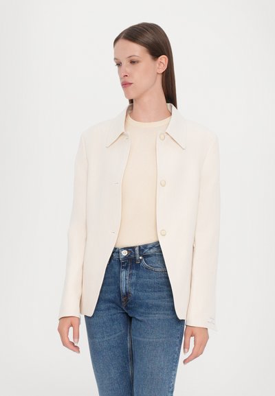 Light beige jacket with a collar, button closure, and long sleeves. Paired with a cream top and blue jeans. Smooth texture, minimalist design.