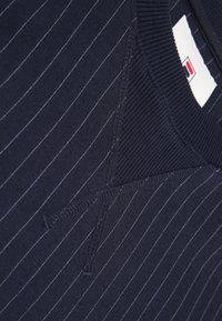 Tessuto blu navy con strisce verticali bianche, caratterizzato da un colletto a coste e un design a V. Etichetta del logo visibile.