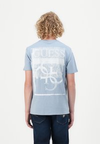 Hellblaue Baumwoll-T-Shirt mit einem großen weißen Grafikdesign, das auf dem Rücken "GUESS" liest. Kurze Ärmel und Rundhalsausschnitt, kombiniert mit dunklen Jeans.