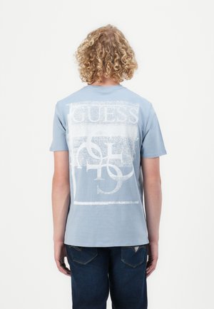 Light blue cotton t-shirt with a large white graphic design reading "GUESS" on the back. Short sleeves and crew neck style, paired with dark jeans.