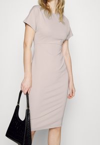 Ljus mauve åtsittande klänning med korta ärmar, rund halsringning och en slimmad silhuett. Modellen håller en svart handväska med krokodilprägling.