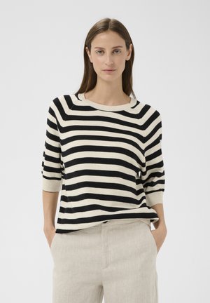 Femme aux longs cheveux bruns portant un pull à rayures noires et blanches et un pantalon beige, les mains dans les poches, debout devant un fond uni.