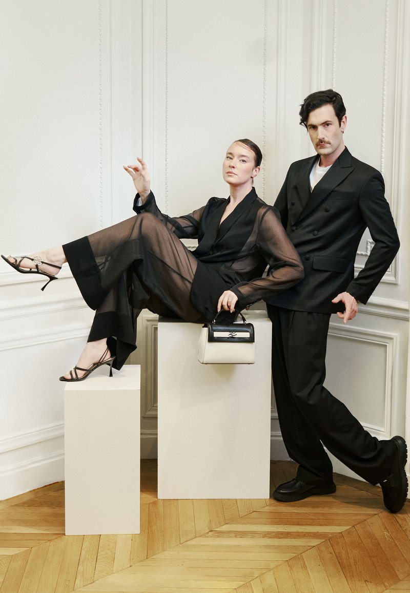 Δύο μοντέλα με μαύρες ενδυμασίες ποζάρουν σε ένα λευκό δωμάτιο· το ένα κάθεται σε βάθρο με τα πόδια τεντωμένα, κρατώντας μια ασπρόμαυρη τσάντα.