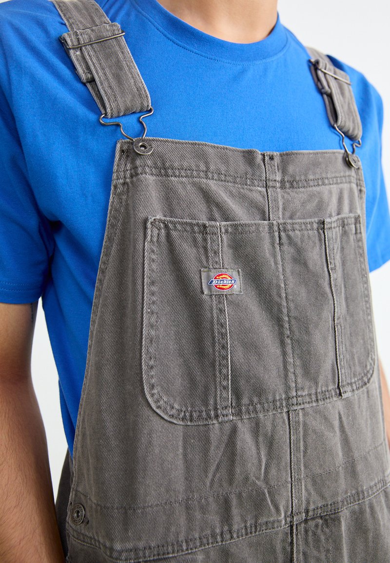 Salopette en denim gris avec des clips en métal, poche avant et logo Dickies ; portée sur une chemise bleue. Le matériau présente une légère texture et des plis.