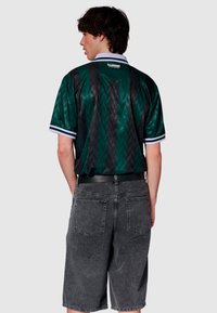 Svart och grön kortärmad skjorta med en chevron-mönster, krage och vita detaljer, kombinerad med gråa jeansshorts. Visar bakifrån.