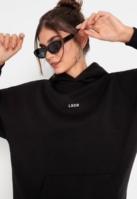 Felpa nera oversize con tasca frontale, caratterizzata da un piccolo logo bianco "LSCN". Indossata con occhiali da sole neri a forma di gatto, che evidenziano una texture liscia.