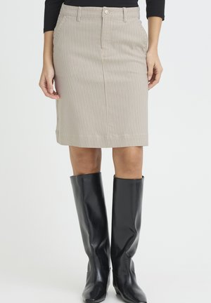 Frau trägt einen knielangen Rock mit beigen vertikalen Streifen, ein schwarzes langärmeliges Oberteil und schwarze kniehohe Stiefel, steht vor einem schlichten Hintergrund.