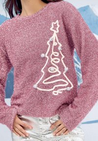 Mirdzoša rozā džemperis ar garām piedurknēm, kurā ir balta Ziemassvētku koka kontūra un pom-pom detaļas, stilizēts virs metāliskām biksēm.