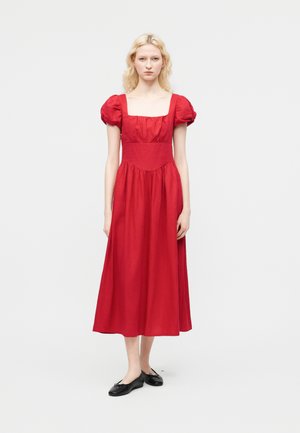 Frau mit heller Haut und blondem Haar trägt auf einfachem Hintergrund ein rotes Midikleid mit kurzen Puffärmeln und schwarze Ballerinas.