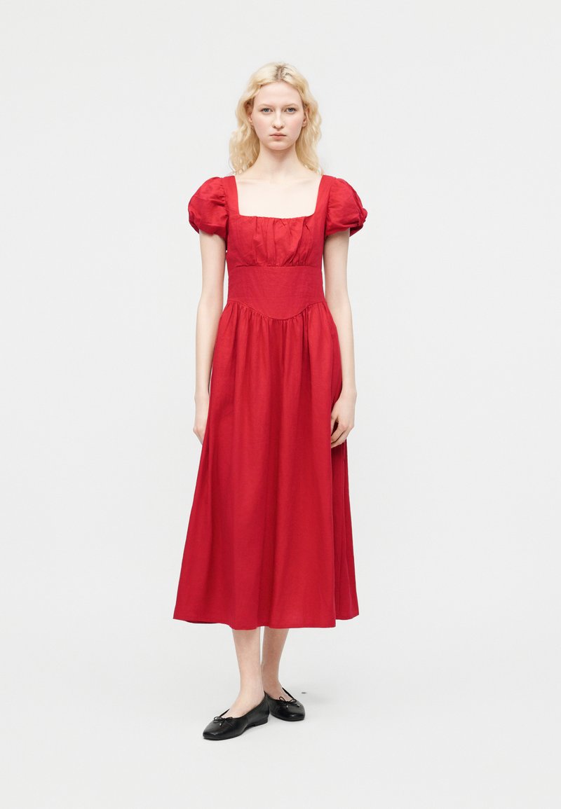 Femme à la peau claire et aux cheveux blonds portant une robe midi rouge à manches courtes bouffantes et des ballerines noires, sur un fond uni.