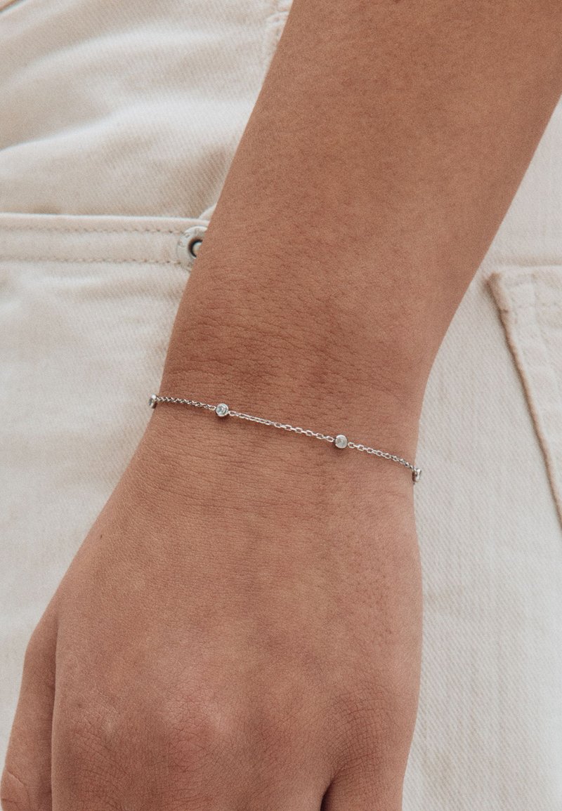 Srebrna bransoletka z łańcucha z małymi okrągłymi akcentami, noszona na nadgarstku, o delikatnym wzorze i gładkiej fakturze na skórze.