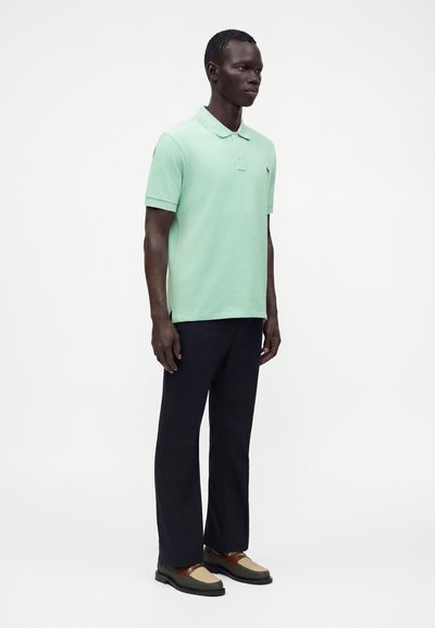 Homme portant un polo à manches courtes vert menthe, un pantalon noir et des chaussures tissées marron et beige, debout devant un fond blanc uni.