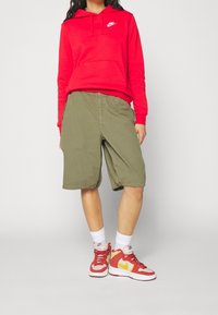 Sweat à capuche rouge avec poche frontale et logo blanc, short vert olive et baskets rouge-jaune. Les chaussettes sont blanches et de style mi-mollet.