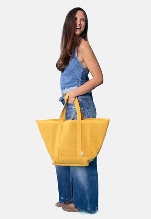 Woman with long brown hair wearing denim top and jeans, smiling while holding a large yellow tote bag by its handles.