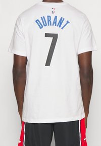 Camiseta de algodón blanca con "DURANT" en azul y el número "7" en malla negra. Logotipo de la NBA encima del nombre, mangas cortas y corte relajado.