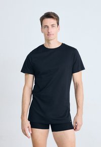 Man standing against white background wearing a plain black t-shirt and black boxer briefs, looking straight at the camera.