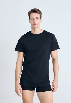 Homme se tenant contre un fond blanc, portant un t-shirt noir uni et un boxer noir, regardant droit vers la caméra.