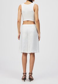Femme debout, portant un crop top blanc sans manches, une jupe blanche jusqu'aux genoux avec des poches arrière, et des talons noirs à brides, sur un fond uni.