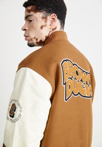 Smeđi i krem bomber jakna s velikim logotipom "BROWN DUCKS" na leđima. Materijal je teksturiran s kontrastnom glatkom rukavom.