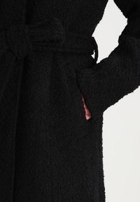 Musta tekstuuri takki, jossa on vyötärölle kiristettävä vyö, suuret sivutaskut ja pehmeä, karvainen kangas. Lähikuva kädestä, joka ojentuu taskuun.