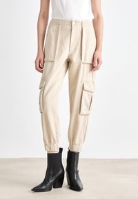 Beige Cargo-Hosen mit elastischen Bündchen, mehreren Seitentaschen und geradem Schnitt. Kombiniert mit schwarzen Knöchelstiefeln mit Blockabsatz.