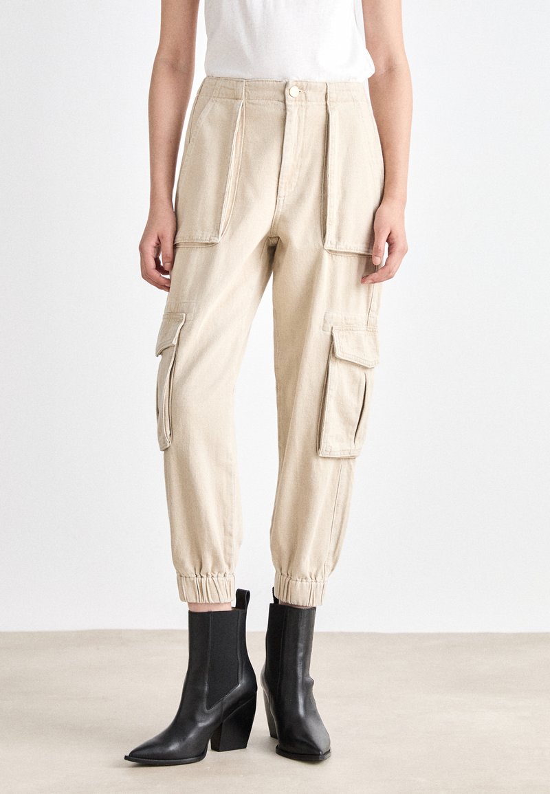 Beige Cargo-Hosen mit elastischen Bündchen, mehreren Seitentaschen und geradem Schnitt. Kombiniert mit schwarzen Knöchelstiefeln mit Blockabsatz.