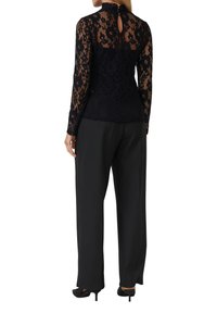 Haut en dentelle noire à manches longues avec un col haut et un dos en clef, associé à un pantalon noir ample et des talons noirs.