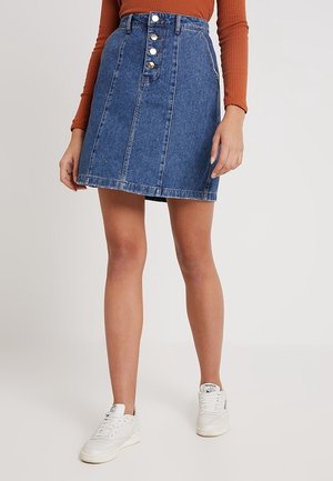 Gonna a trapezio in denim, blu, con cinque bottoni frontali, orlo sfrangiato, abbinata a una maglia a maniche lunghe a coste e sneakers bianche.