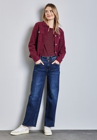 Burgunderfarbene Knopfjacke über einem kastanienbraunen Oberteil, kombiniert mit weit geschnittenen, dunklen Jeans und weißen Turnschuhen. Einfaches Design, lässiger Look.