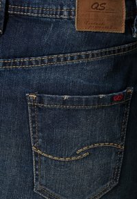 Pantalones vaqueros de mezclilla en índigo oscuro con costuras amarillas contrastantes. Presentan un bolsillo trasero con un pequeño logo "QS" rojo y una etiqueta de cuero.