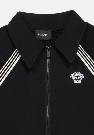 Mustast lukuga jakk kraega, valgete küljeribadega ja valge tikitud Medusa pea logoga rinnal Versace sildi all.