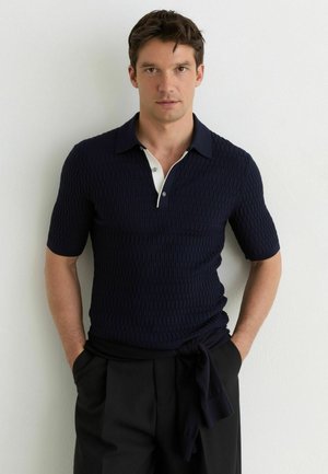 Bărbat purtând un tricou polo texturat bleu închis cu mâneci scurte, cu pată de nasturi albași și pantaloni negri, stând lipit de un perete alb cu mâinile în buzunare.