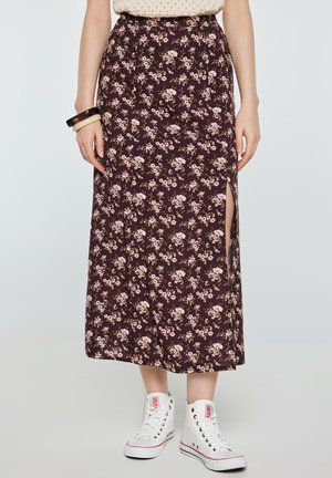 Kobieta ubrana w fioletową kwiecistą spódnicę midi z bocznym rozcięciem, w połączeniu z białymi wysokimi trampkami i dwoma bransoletkami na lewej ręce.