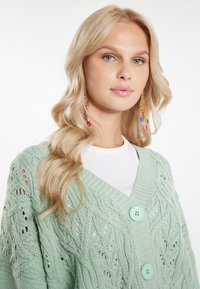 Mintgrön kabelstickad cardigan med V-ringning, med stora knappar och en strukturerad mönster. Bärs över en vit T-shirt.