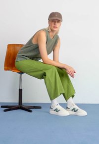 Grüne Cordhose mit weitem Bein, graues strickloses Oberteil, graue Mütze, weiße Sneakers mit grünen Akzenten, sitzend auf einem Holzstuhl vor einem schlichten Hintergrund.