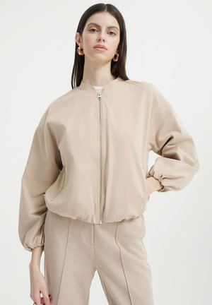 Lehká béžová bomber bunda se strukturovaným designem, elastickými manžetami a předním zipem; obsahuje boční kapsy a hladkou texturu.