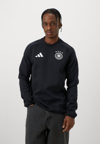 Muškarac s pletenom kosom nosi crnu duksericu dugih rukava s Adidas logom i logom njemačke nogometne reprezentacije te tamne traperice, stoji nasuprot običnoj pozadini.